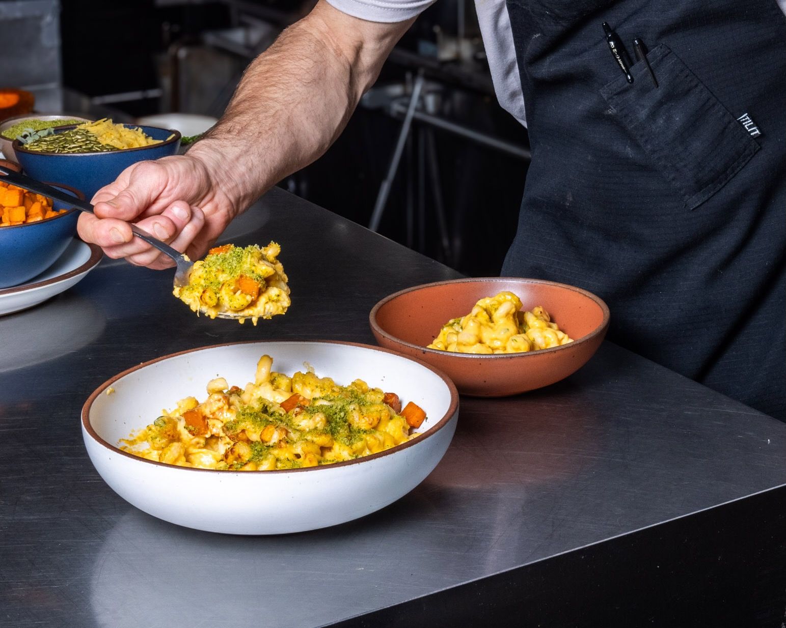 In a commercial kitchen, two ceramic bowls filled with a gourmet mac and cheese with a hand plating a spoonful into one of the bowls.