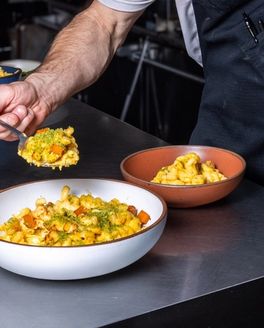 In a commercial kitchen, two ceramic bowls filled with a gourmet mac and cheese with a hand plating a spoonful into one of the bowls.