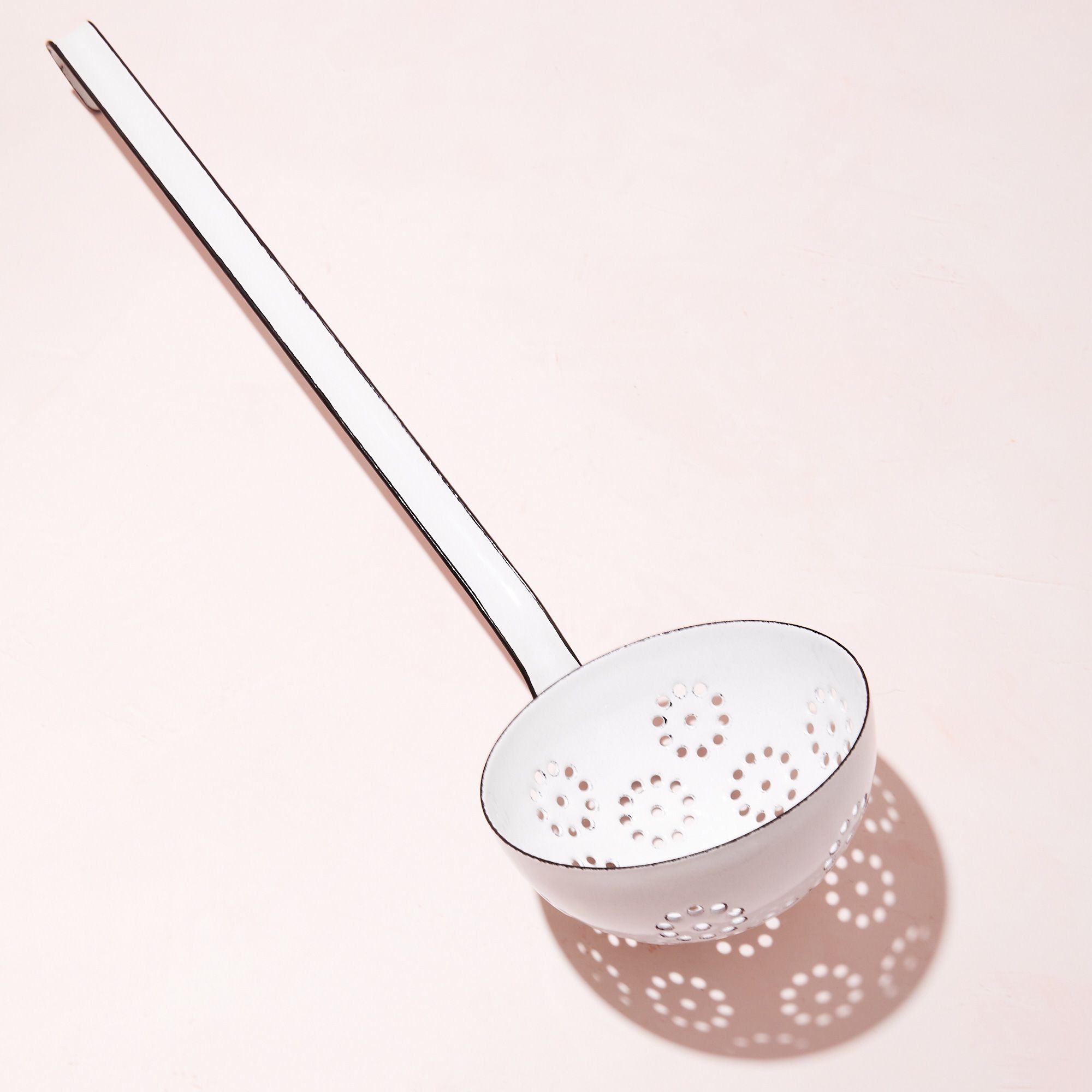 A white enamel ladle with little slotted holes inside the ladle scoop and its shadow on a soft pink background