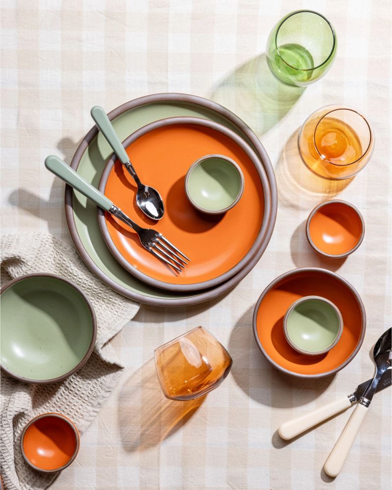 An overhead view of ceramic plates and bowls artfully arranged in bold orange and sage green colors, surrounded by flatware glasses and more. All against a natural gingham tablecloth.