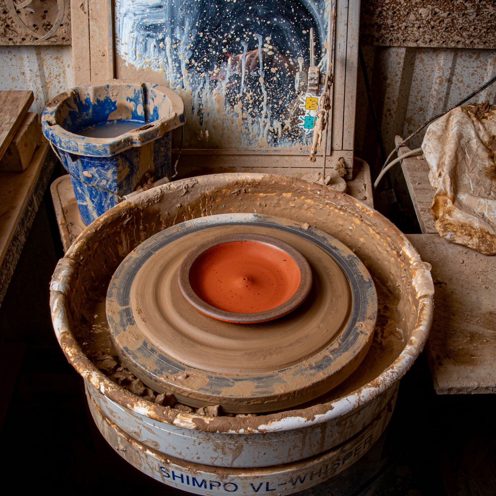 In a pottery studio setting and on a potter's wheel sits an orange plate-shaped incense holder with an unglazed rim featuring iron speckles.