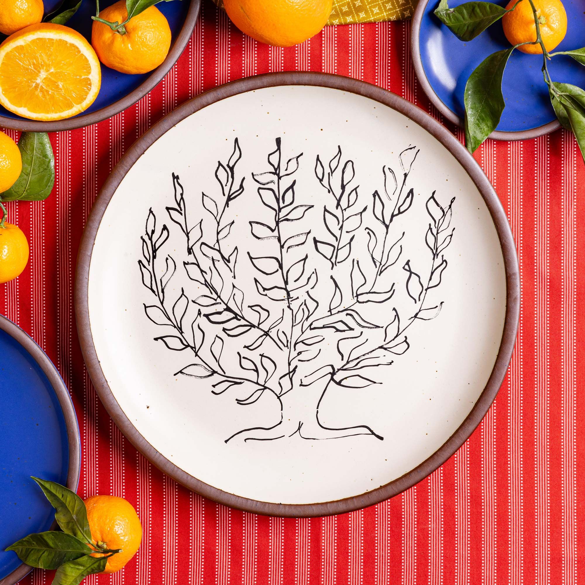 A large serving platter in a warm off-white color with unglazed rim with a black decal of the artwork, Le Platane, from Henri Matisse, sitting on a red tablecloth near cobalt blue plates and oranges.