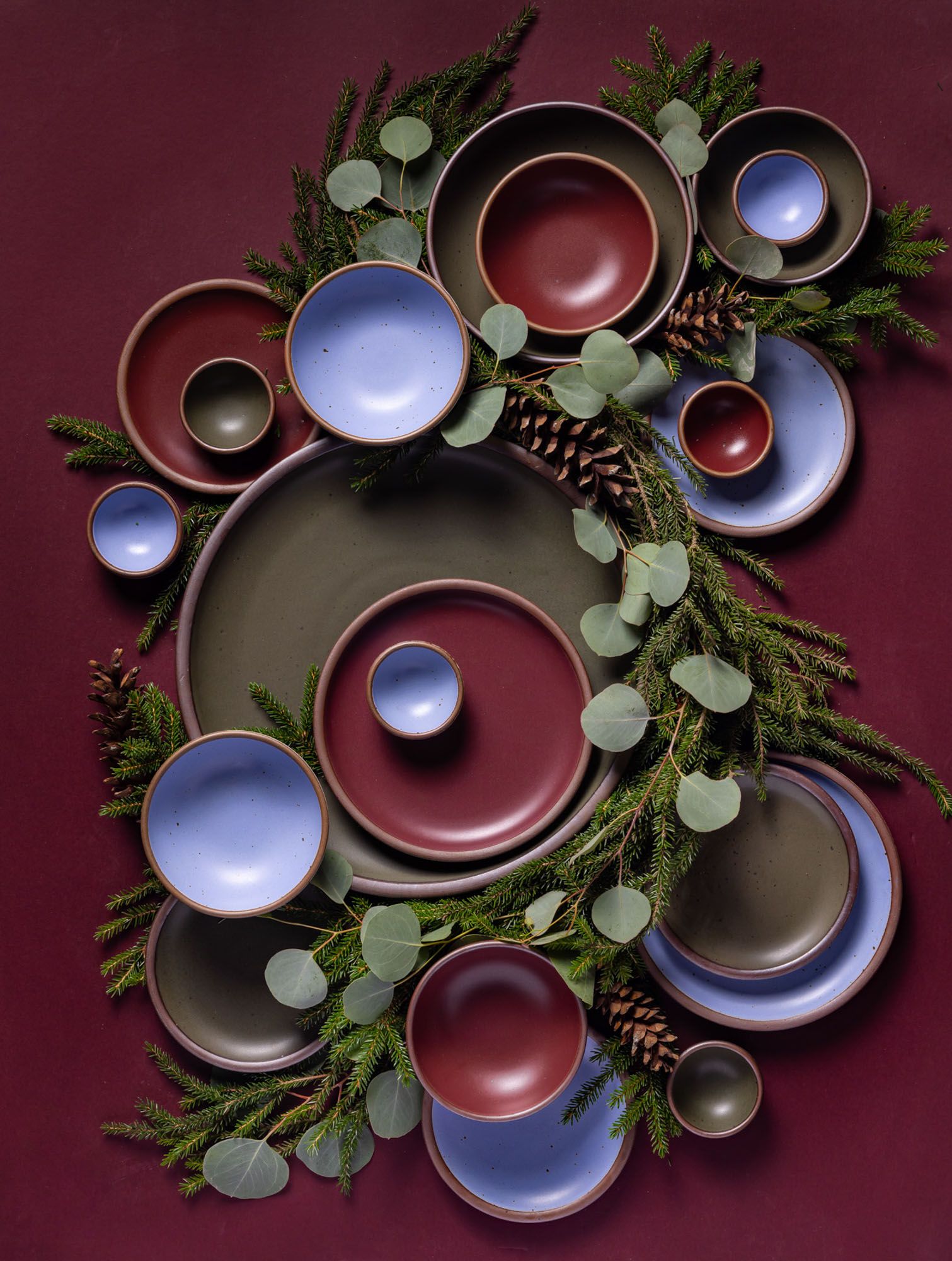 An artful arrangement of ceramic plates and bowls in various sizes in plum, dark forest green, and periwinkle colors, surrounded by pine cones and eucalyptus.