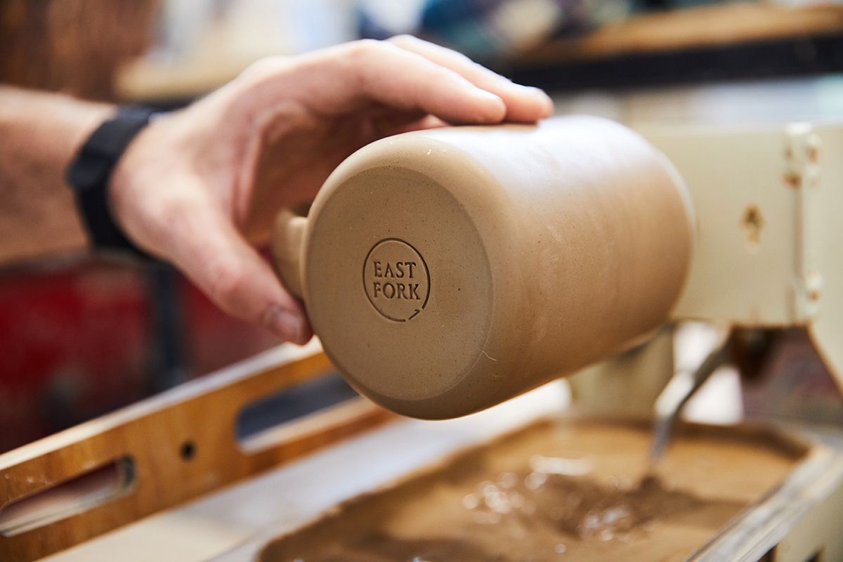 A mug in the process of being made, a hand holds in while it is sitting on a machine with a fresh stamp on the bottom that reads "East Fork"