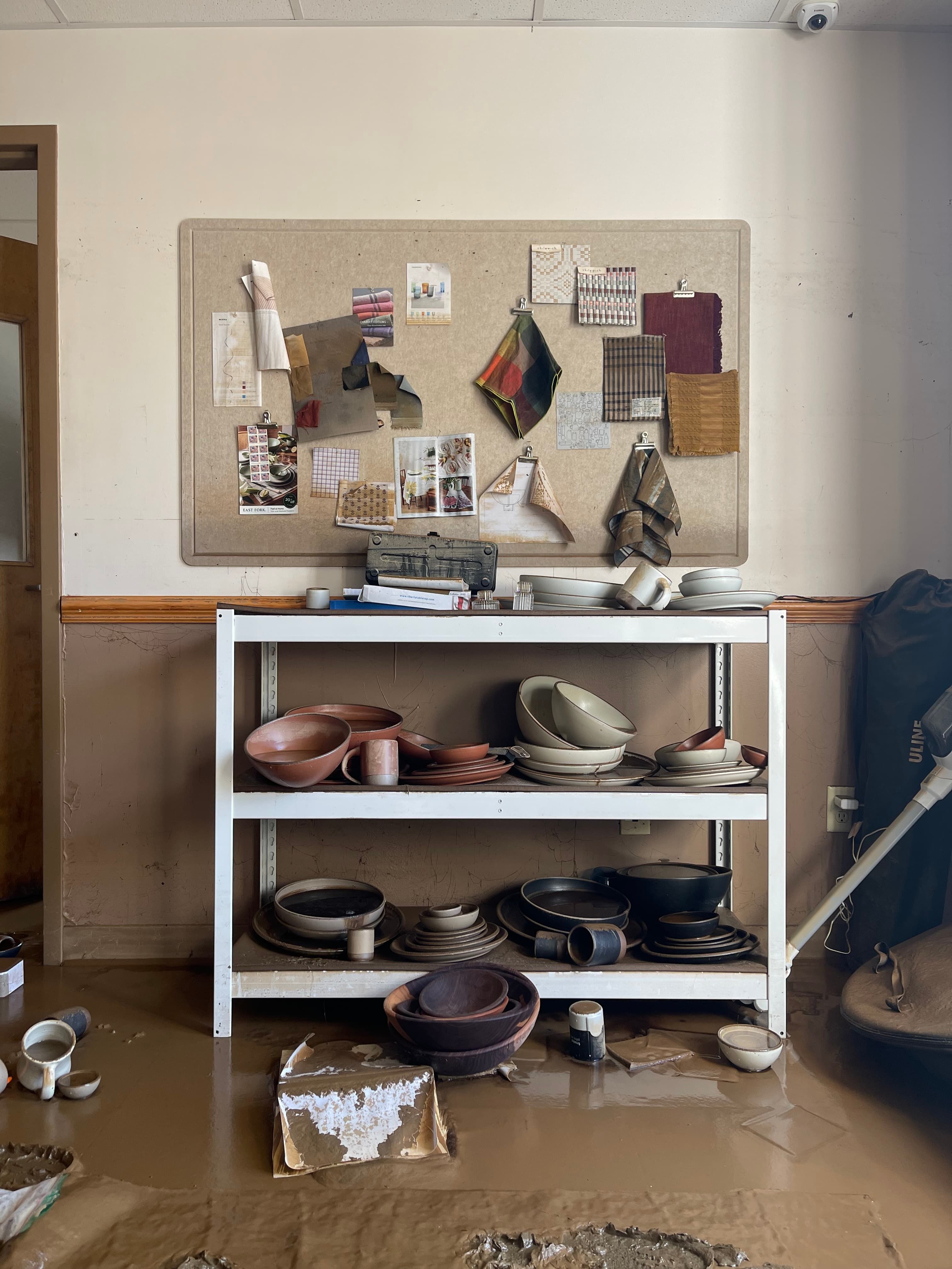 A shelf with pottery and a mood board above it are damaged by the storm, with mud all over the floor.