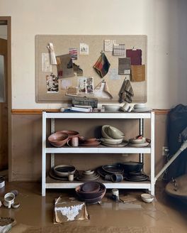 A shelf with pottery and a mood board above it are damaged by the storm, with mud all over the floor.