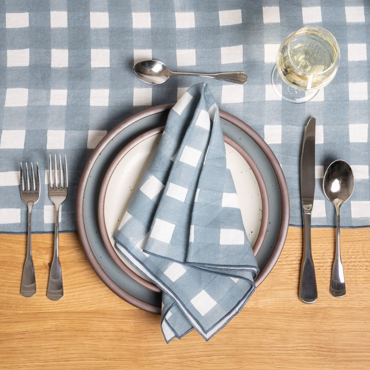 A stainless steel five-piece flatware including 2 forks, 2 spoons, and 1 knife, in a polished finish and classic design. This set is on a place setting with ceramic dinnerware, glass, and blue-grey crosshatch napkin and table runner.