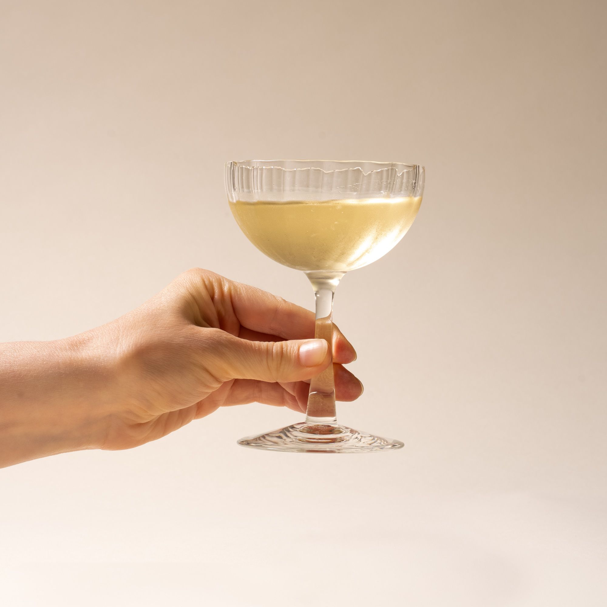 A hand holds out white wine in a clear ribbed glass coupe with a tall stem casting a soft shadow on a beige background.
