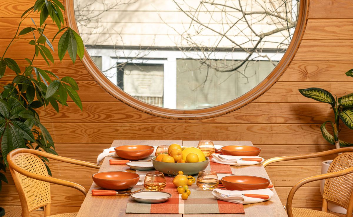 A table set with a gingham table runner, bold orange bowls and cool white plates, orange-handled flatware, whiskey snifters, and a centerpiece of fruits and vegetables. Also pictured is a woven chairs at the table table, a large houseplant, and a large circular window.