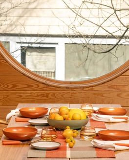 A table set with a gingham table runner, bold orange bowls and cool white plates, orange-handled flatware, whiskey snifters, and a centerpiece of fruits and vegetables. Also pictured is a woven chairs at the table table, a large houseplant, and a large circular window.