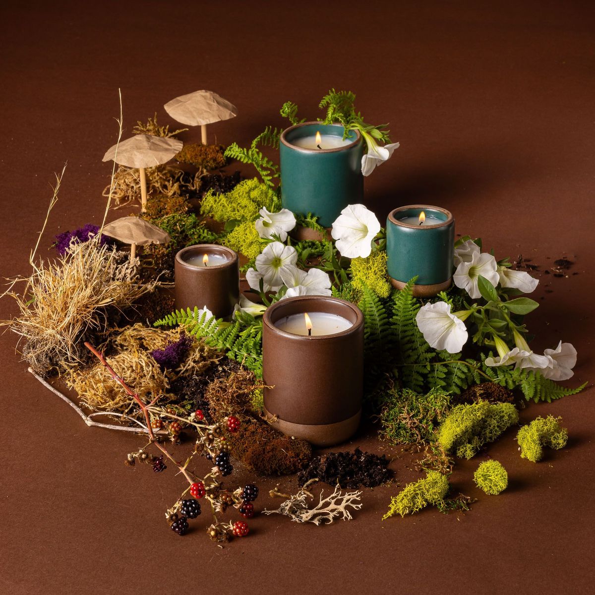 Four candles in ceramic vessels in dark teal and brown colors. They are artfully arranged and surrounded by moss, flowers, dried grass, mushrooms, and berries.