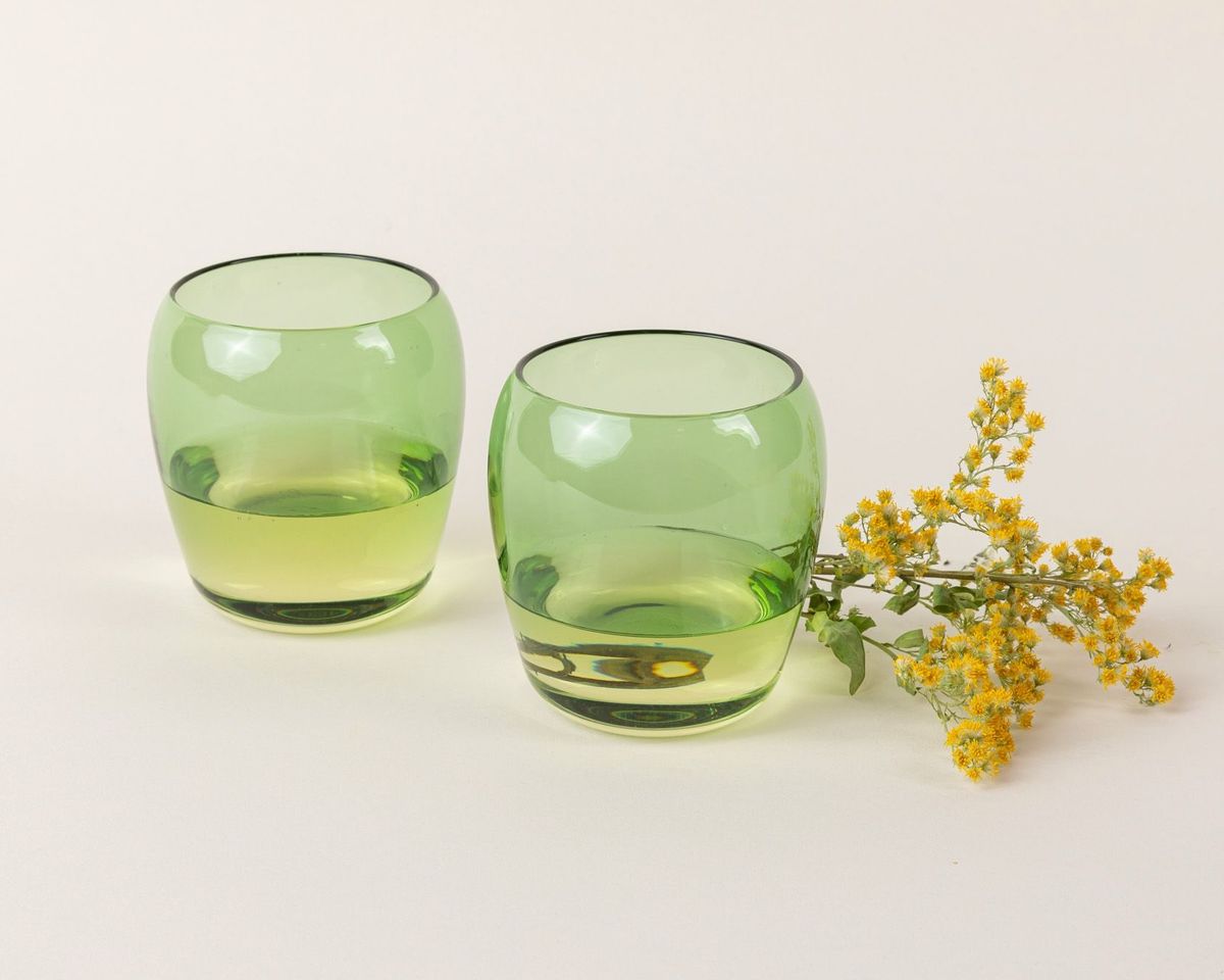 Two short light greenglass tumblers with a slight curve inward on the top. The glasses are filled with white wine and a flower is sitting behind them.