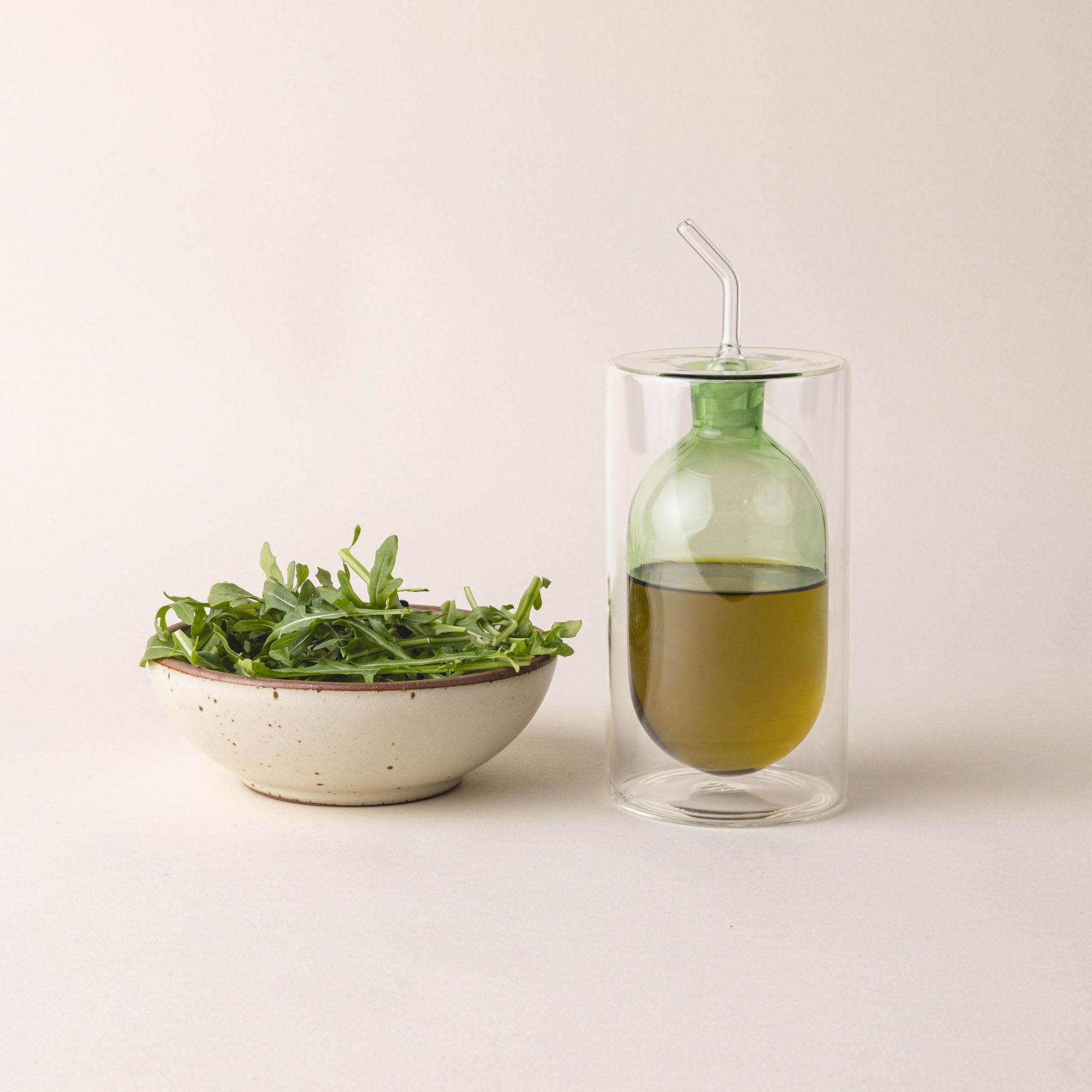 An artful glass cruet in a green color and next to a small side salad. There is a rounded bulb that acts as a container for the cruet, surrounded by a doubled glass wall. The cruet is filled with oil.