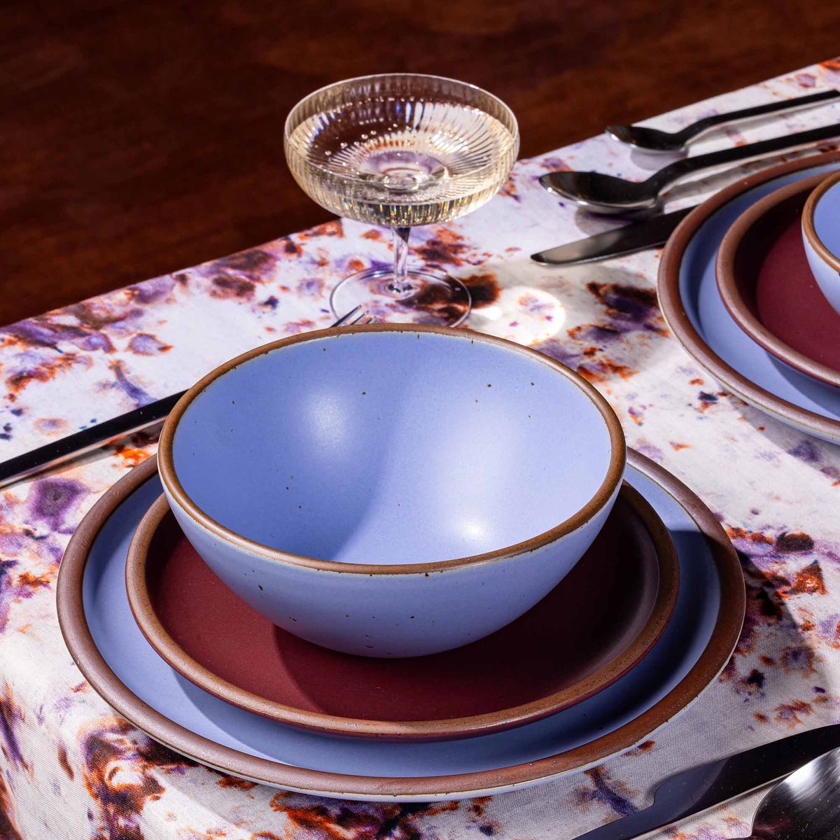 A table setting featuring marbled textiles, a coupe glass, and dishes in plum and periwinkle colors