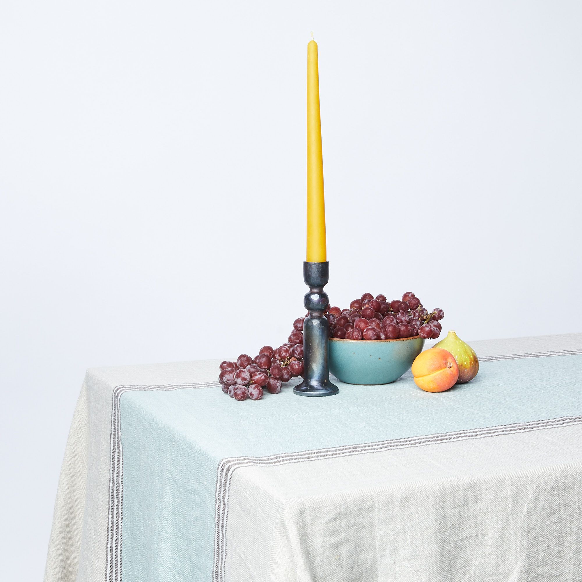 White linen tablecloth with aqua stripe down the middle draped over table with candlestick and fruit bowl on top