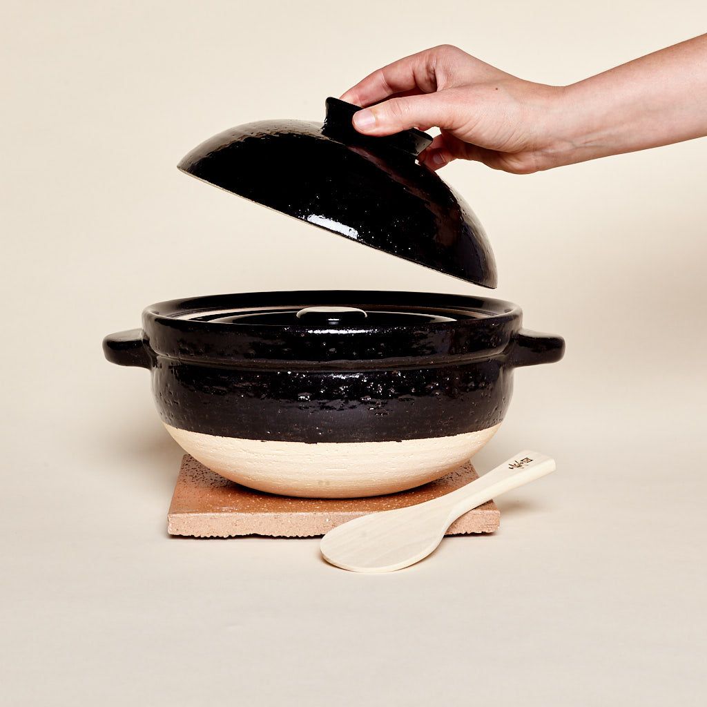 A black-and-neutral color claypot with handles and a shallow-dome shaped lid. The lid is being lifted off by a hand. The cooking vessel sits atop a wooden board next to a wooden spoon.