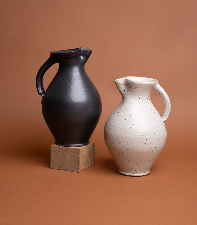 Two sturdy ceramic pitchers against a brown background, one pitcher is glazed in an iron-speckled black, the other in an iron-speckled white.