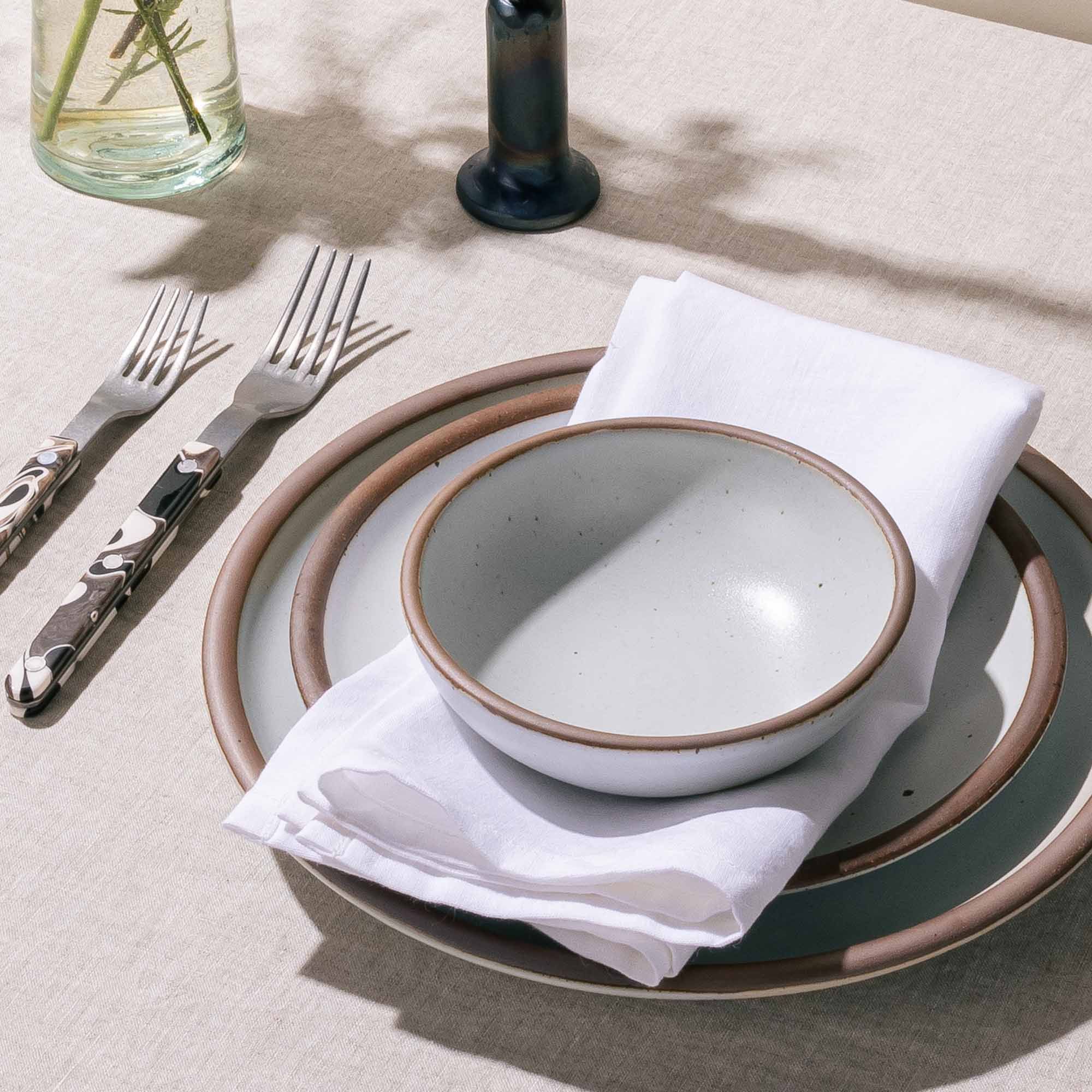 A table setting featuring a cool white dinner plate, side plate, and shallow small bowl with a white napkin with flatware nearby.