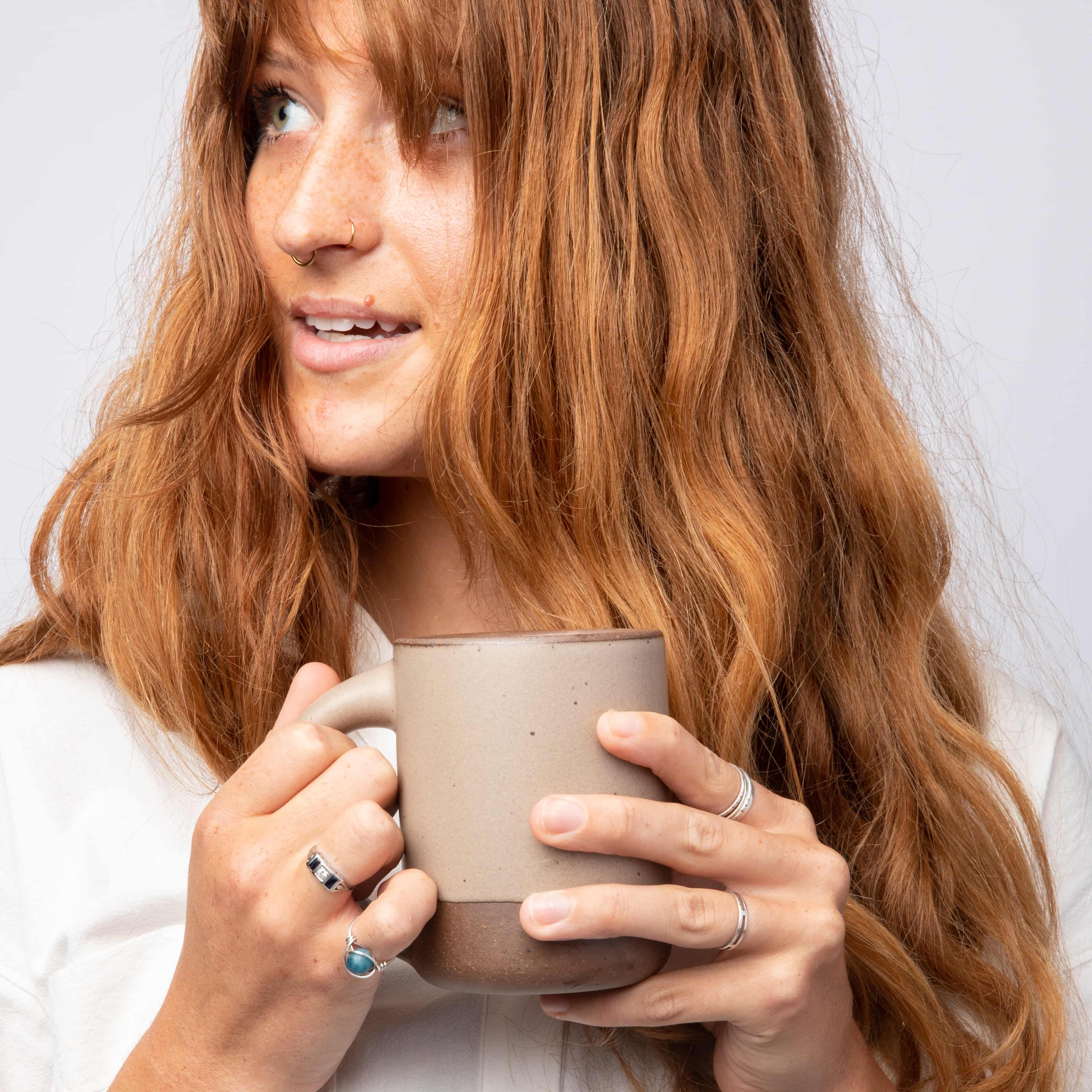 A person with long red hair is holding an East Fork mug in Morel with two hands and looking off to the side.