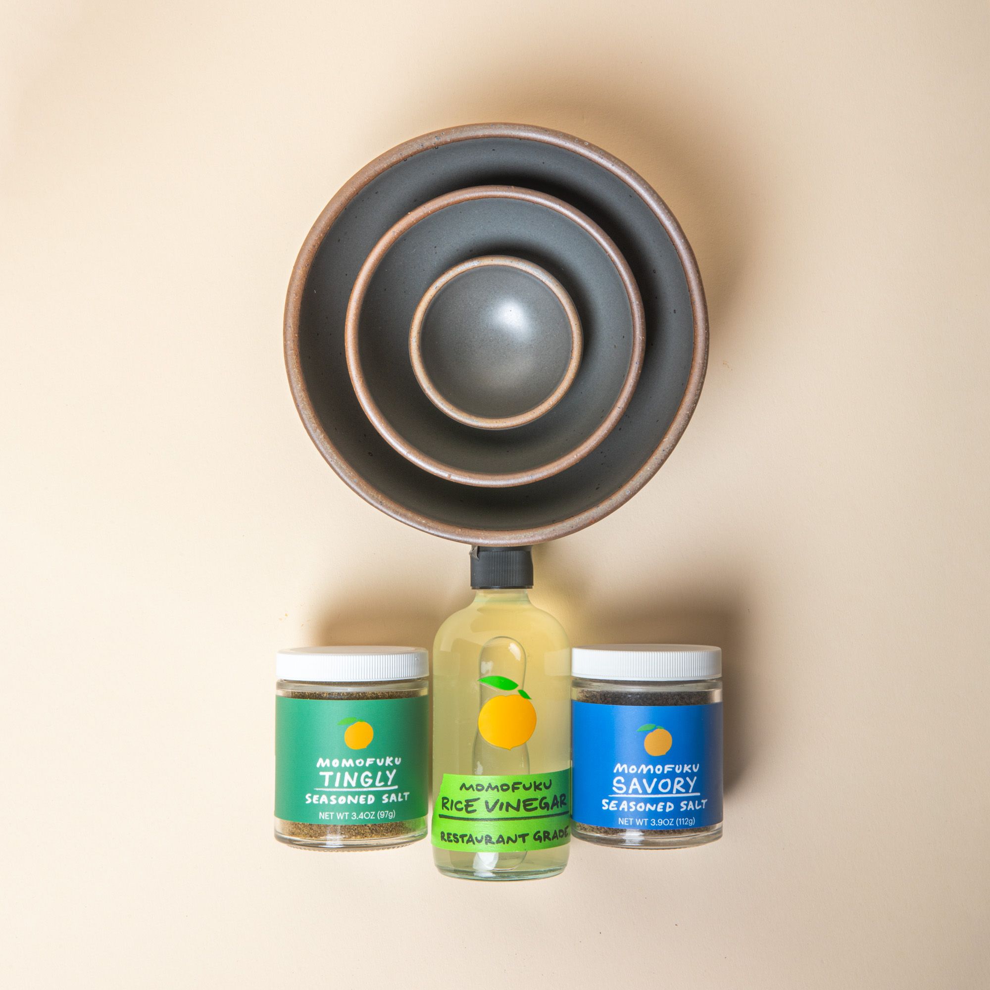 3 Ceramic bowls of varying sizes nested together in a soft cool grey color with 2 jars of Momofuku seasoned salt and a bottle of rice vinegar below.