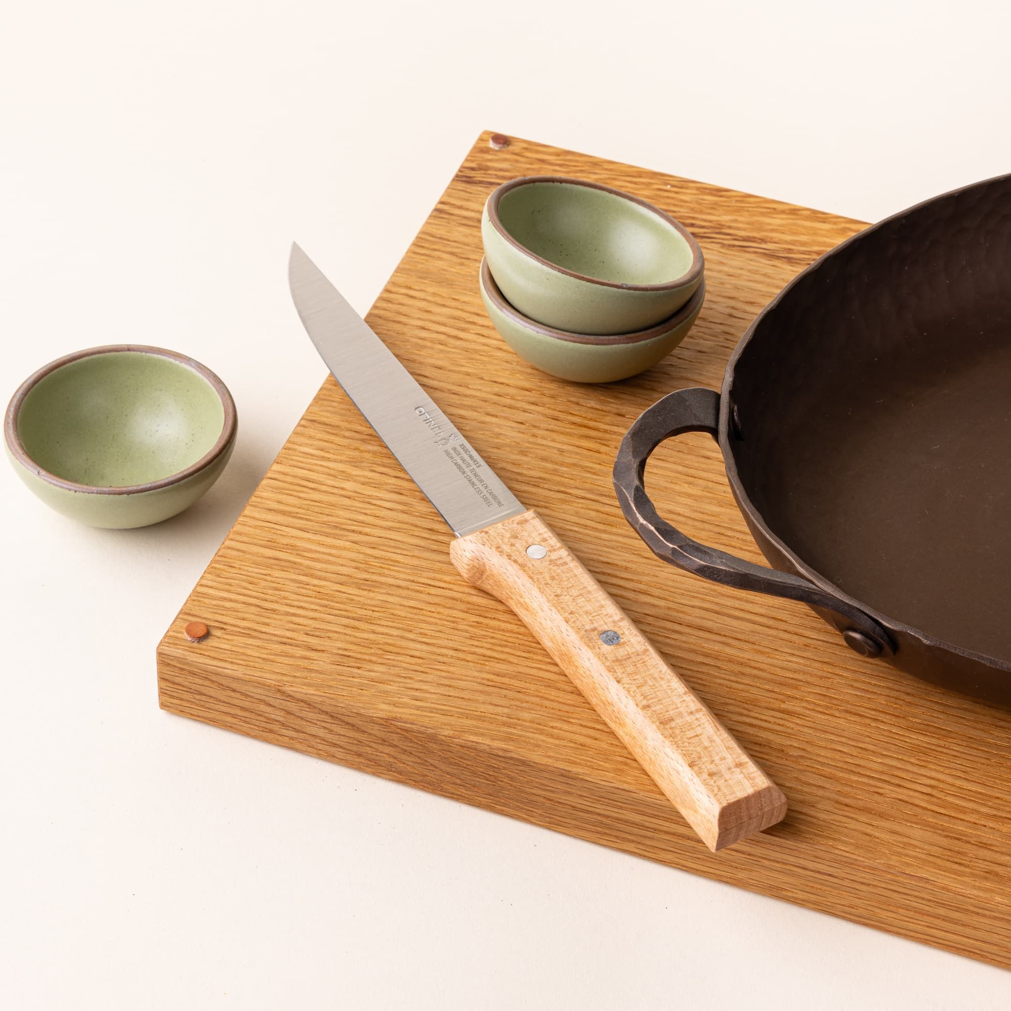 A carving knife with a stainless steel blade and wooden handle sits on a wood cutting board surrounded by an oven roaster and mini ceramic sage bowls.