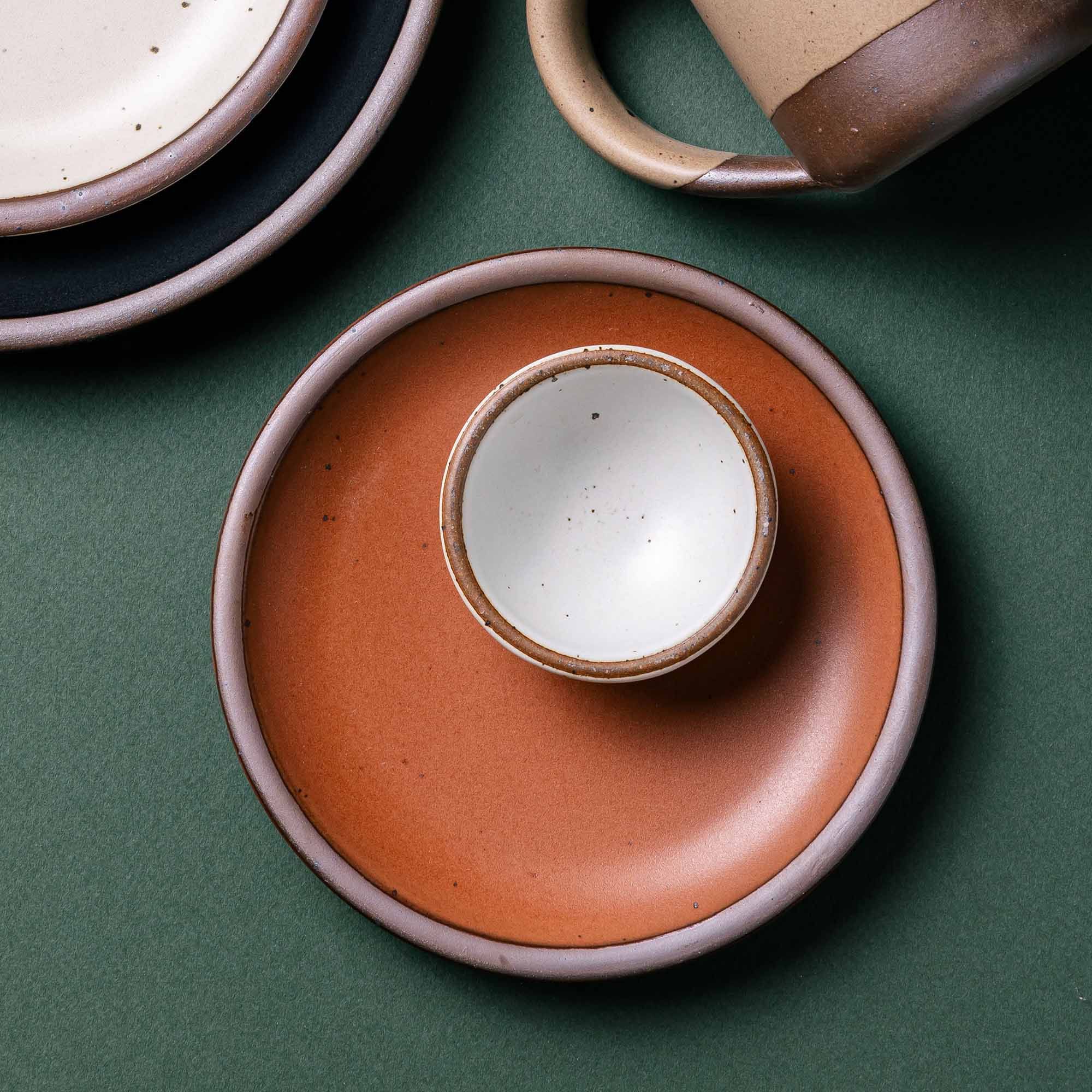 A tiny ceramic bowl in a cool white color sits on top of a dessert sized plate in a burnt terracotta color