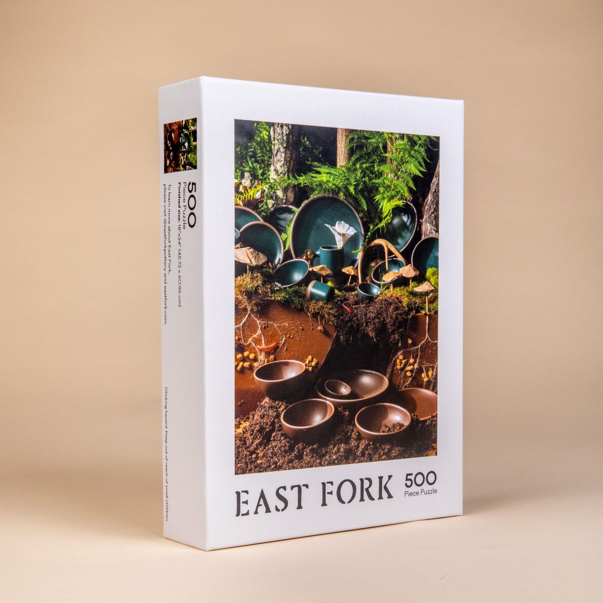 Angled side view of a box of a 500-piece jigsaw puzzle that when put together shows ceramic bowls, plates and mugs in dark teal and brown colors in a natural forest setting.