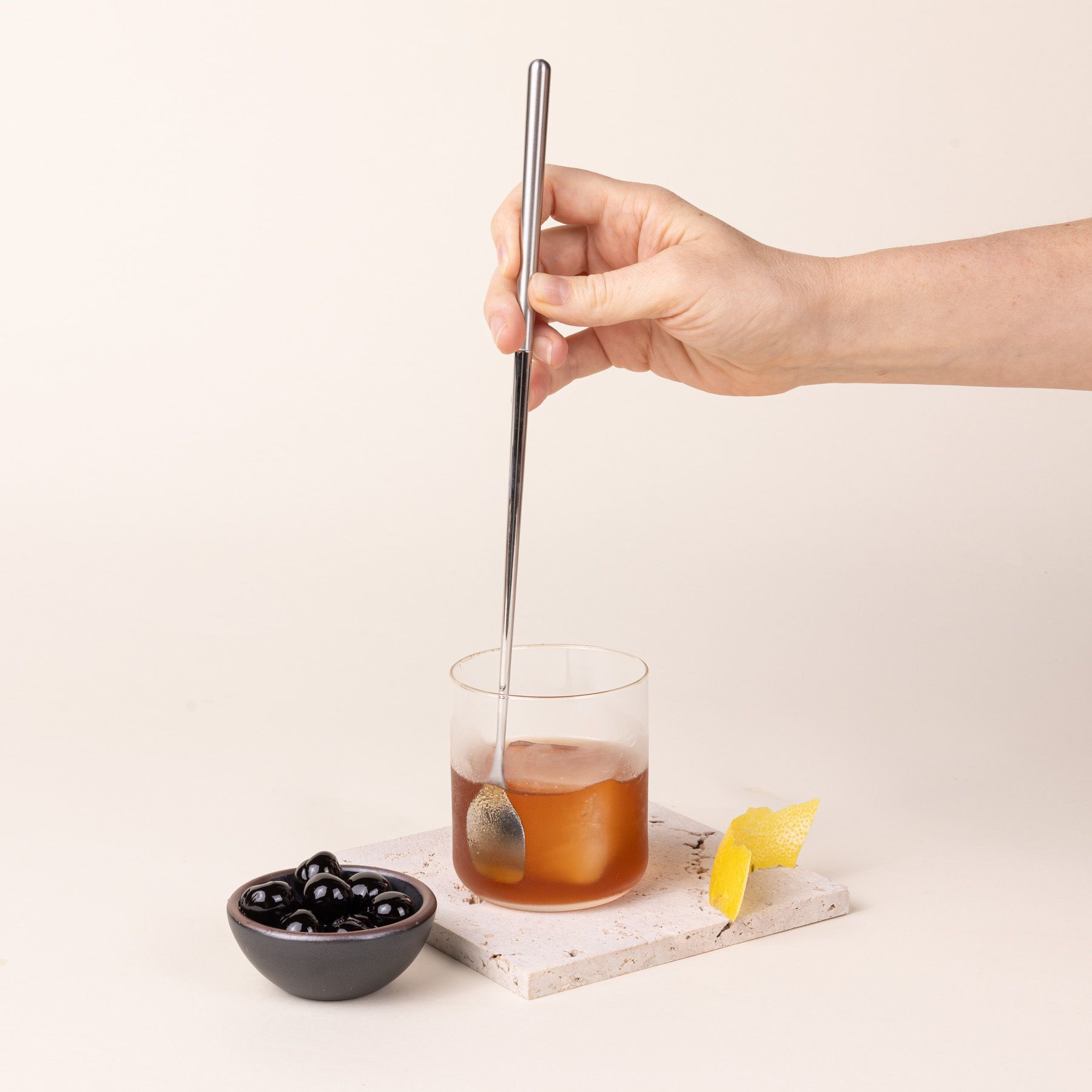 A hand holding a stainless steel spoon stirring a cocktail with a small bowl of cherries nearby