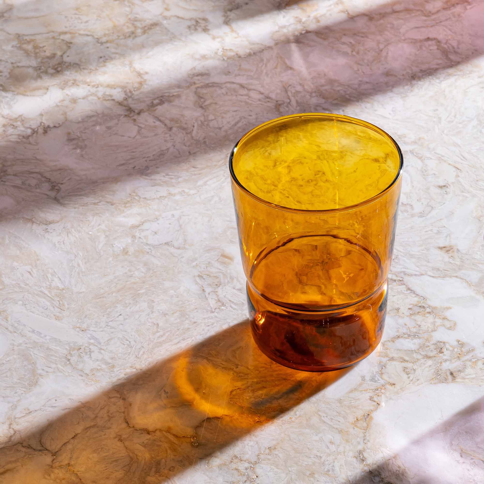 A tumbler glass that widens at the top in an amber color on a table with its shadow casting