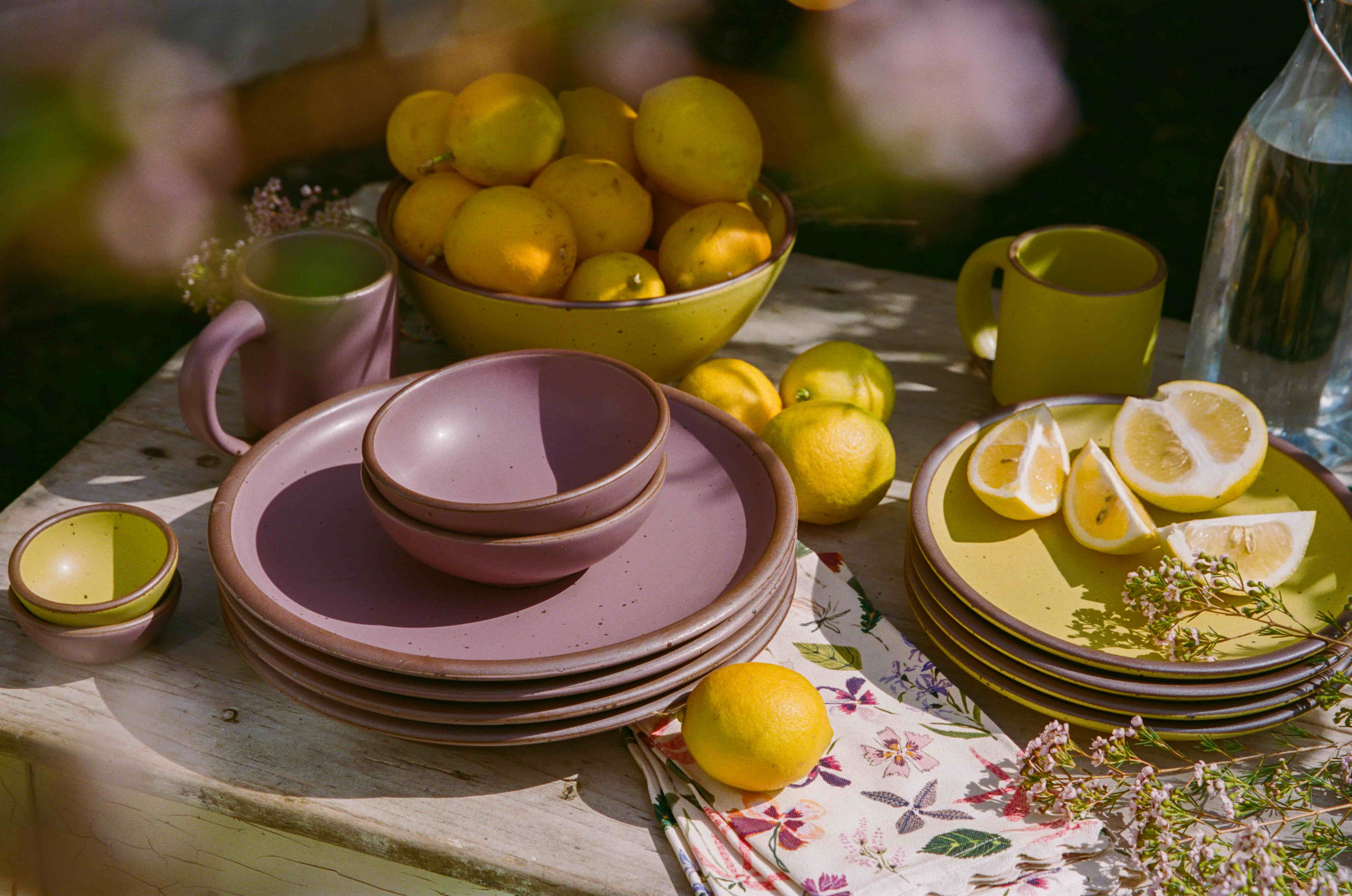 An outside table filled with ceramic dinnerware in zesty yellow and soft mauve purple colors, lemons, floral napkins, and flowers.