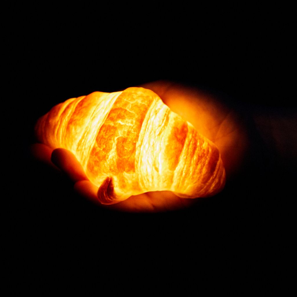 A lamp with the shape, coloring and size of a croissant is illuminated from within in a dark room, where it is held by a hand.