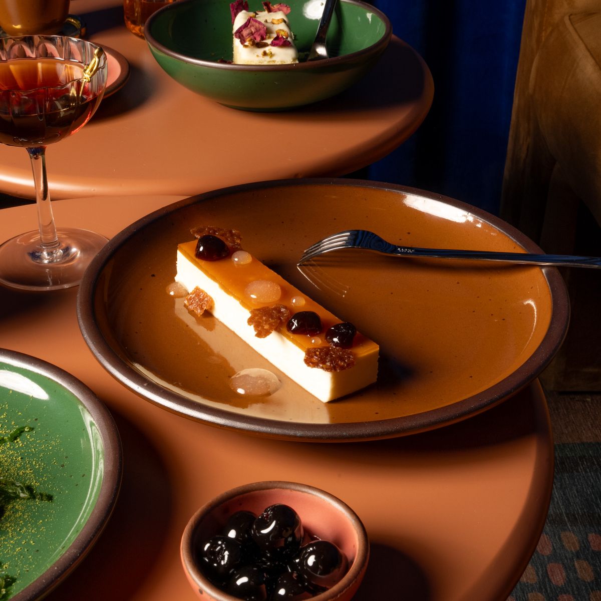 An artful dessert with a fork on a dinner sized ceramic plate in a glossy caramel brown color on a table with ceramic plates and bowls in glossy green and watermelon pink.