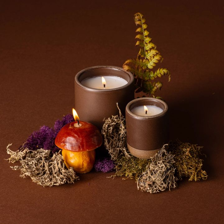 Two lit candles in ceramic vessels in a dark brown color and an artful mushroom candle. They are arranged and surrounded by dried grass and fern leaves.