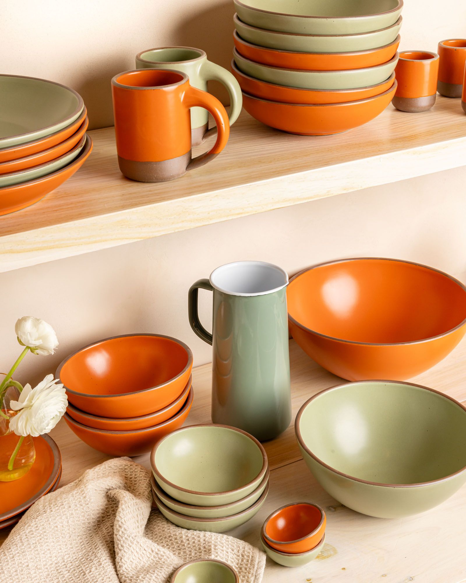 In a kitchen, stacks of ceramic pottery in bold orange and sage green colors, surrounded by a sage green pitcher, flowers, and a kitchen towel.