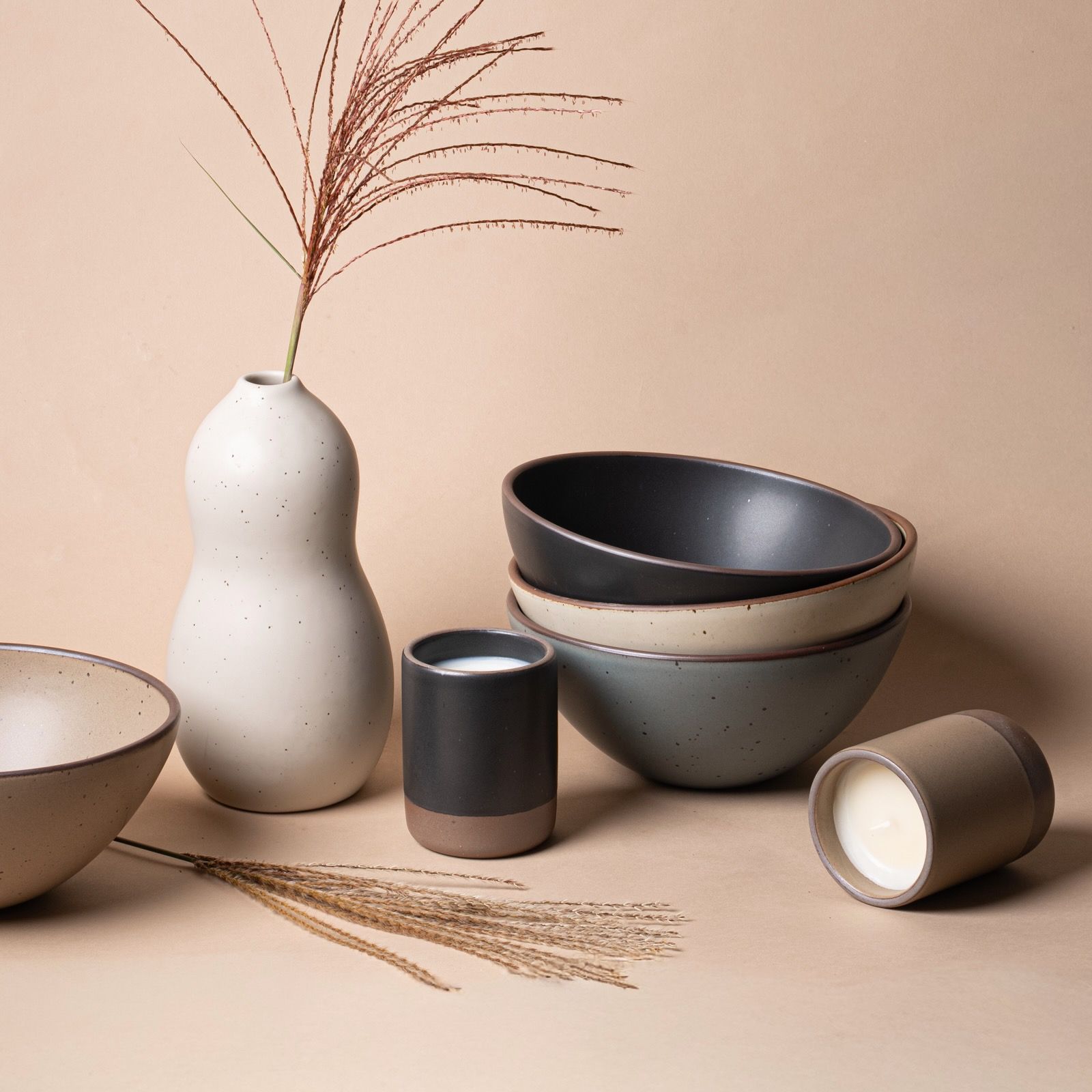 Neutral-toned ceramic bowls, vases, and candles arranged on a beige surface with dried grass for a minimalist, earthy look.