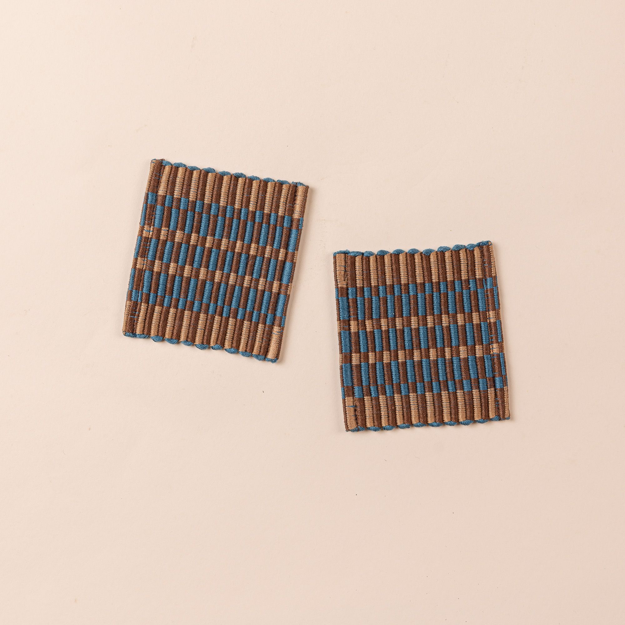 Two square coasters that are hand woven in a striped rectangular grid design in brown, tan, and blue colors