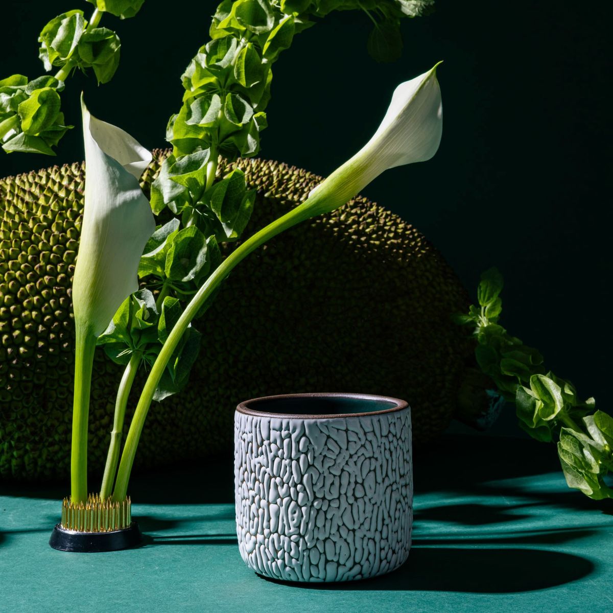 A white ceramic vessel with cracked texture sits next to a gold flower frog with calla lilies