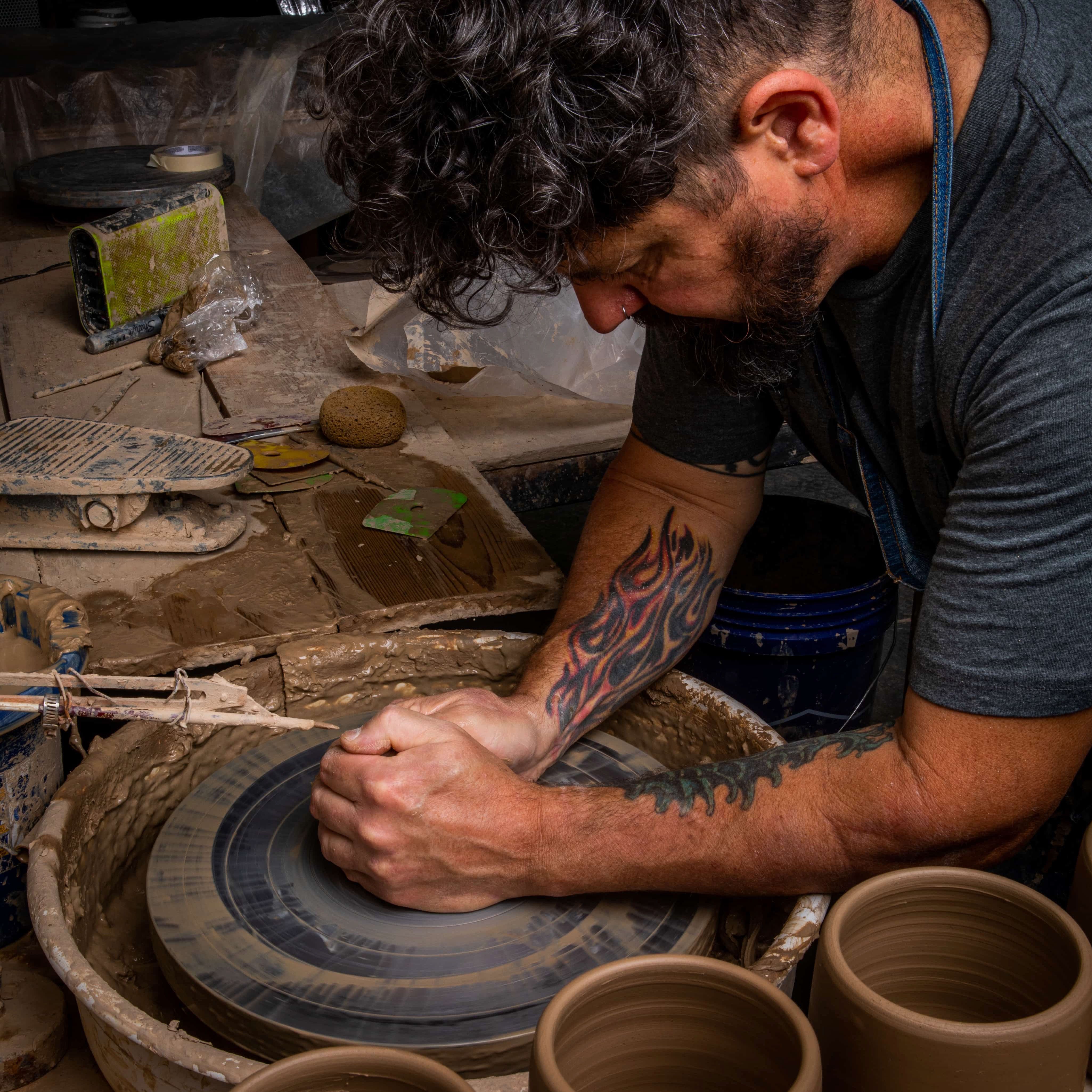 A side profile of a man throwing a ceramic mug on a potter's wheel in a clay workshop environment.