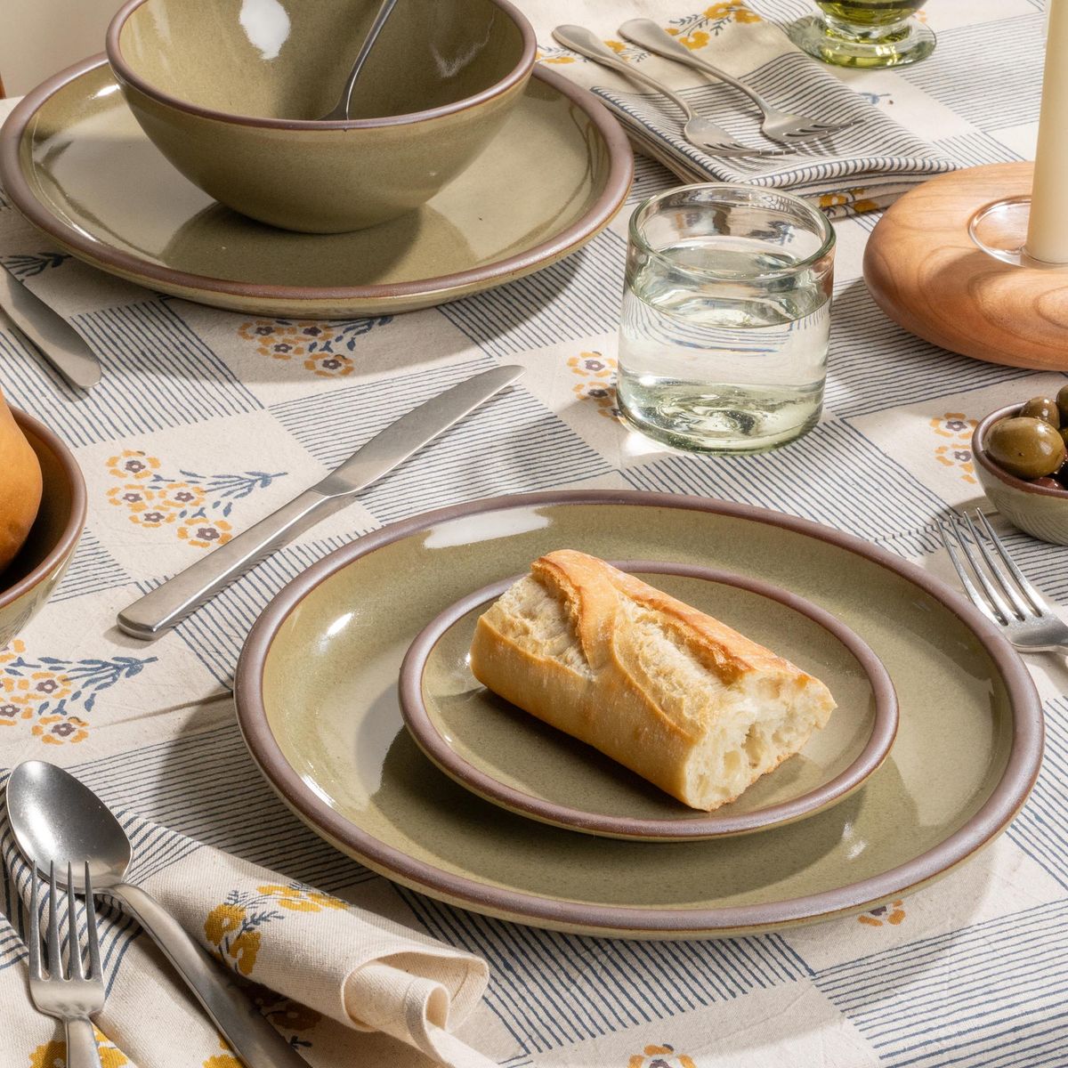 Grey-green stoneware place setting of cake plate and side plate with bread, glass of water, and flatware on a striped floral tablecloth.
