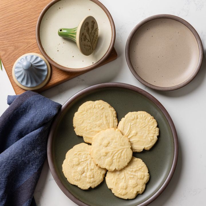 Cookies with a flower stamp are plated on a forest green plate with cookie stamps off to the side with more plates and a blue tea towel.