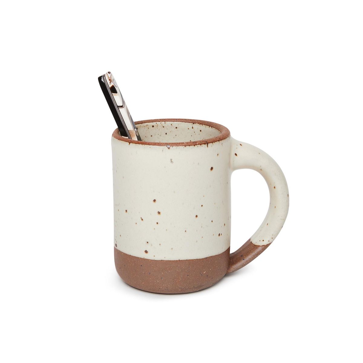 A spoon sitting in a medium sized ceramic mug with handle in a warm, tan-toned, off-white color featuring iron speckles and unglazed rim and bottom base.