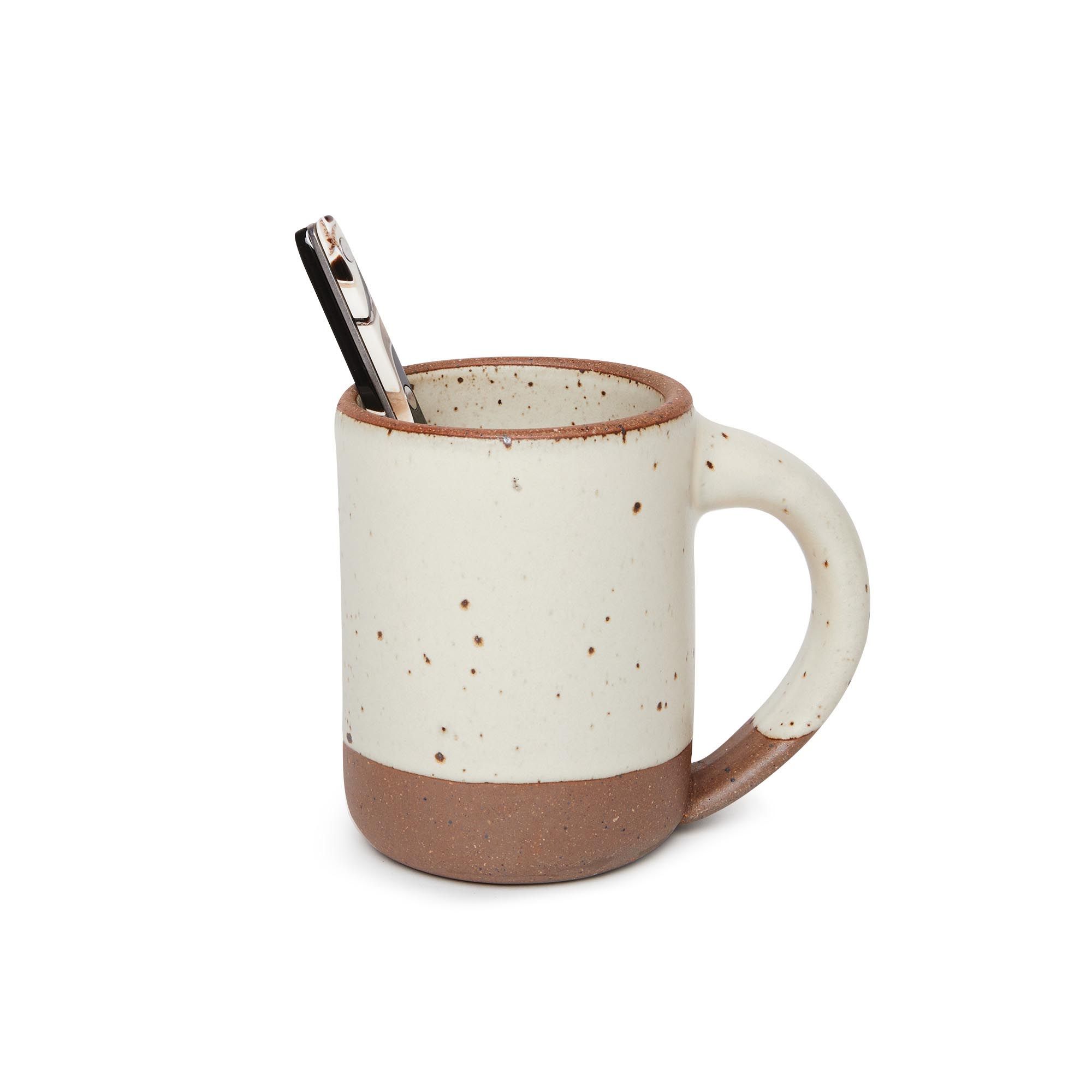 A spoon inside a medium sized ceramic mug with handle in a warm, tan-toned, off-white color featuring iron speckles and unglazed rim and bottom base.