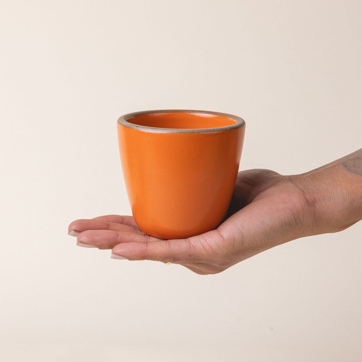 A hand holds a short cup that tapers out to get wider at the top in a bold orange color featuring iron speckles.