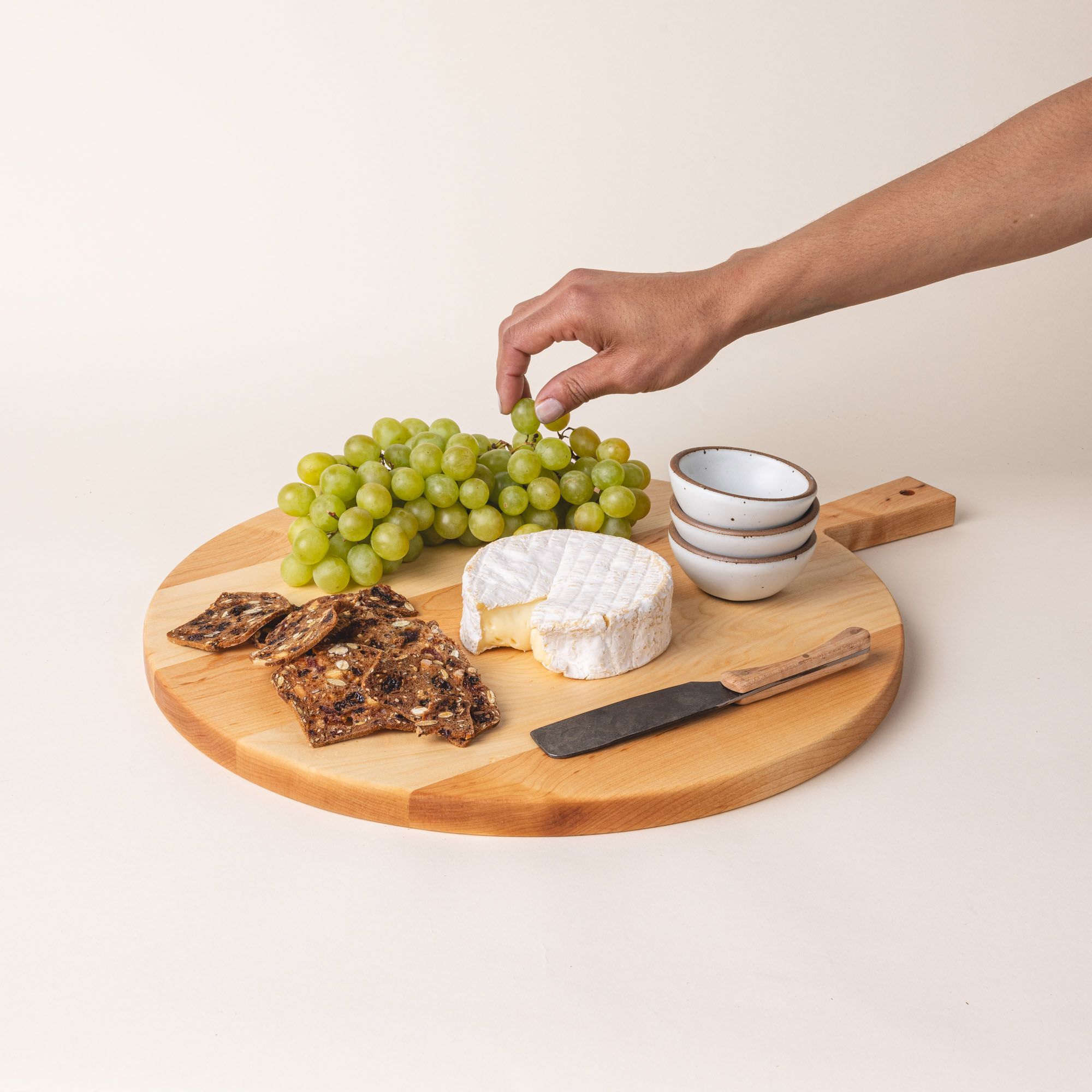 A simple charcuterie board with cheese, crackers, and green grapes is on a round wooden serving board with a short rectangular handle with a hole for hanging