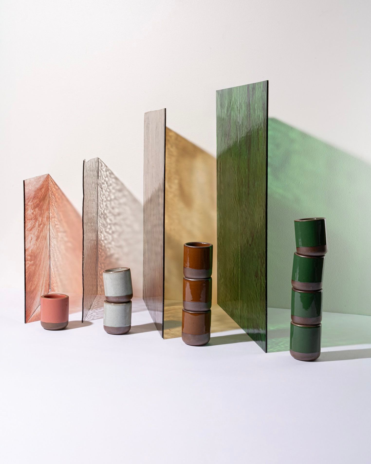 Ceramic cups in shades of pink, icy white, brown, and green are stacked beside vertical textured glass panels casting colorful shadows.