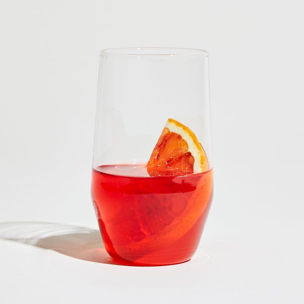 A tall stemless wine glass that taper toward the base, filled halfway with a vermillion liquid and a slice of an orange