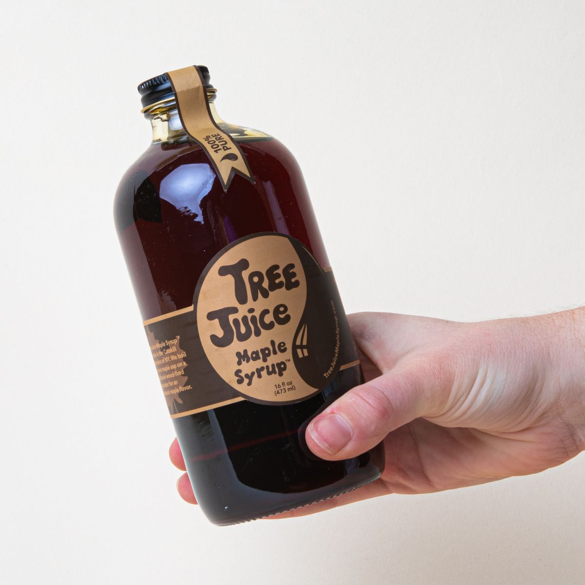 Hand holding a glass bottle with black cap filled with brown syrup and a label that reads "Tree Juice Maple Syrup"