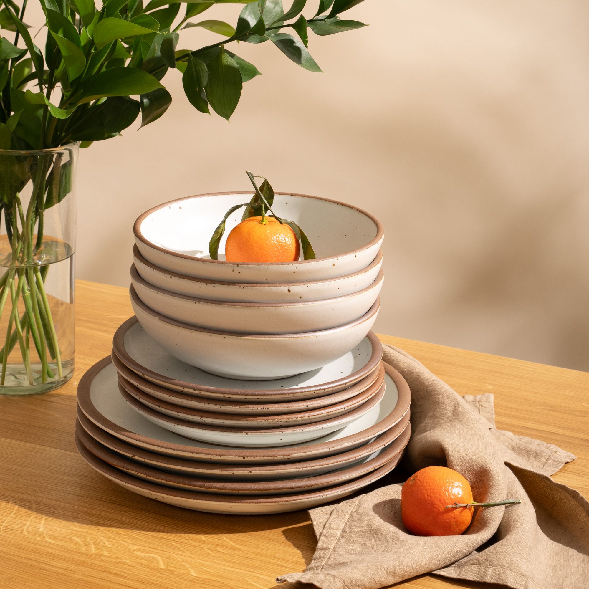 4 ceramic dinner plates, 4 side plates, and 4 shallow dinner bowls in a cool white color with iron speckles and unglazed rims are stacked all together on a table with oranges.