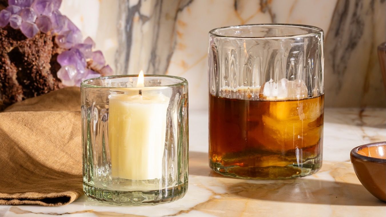 Glass tumbler with a lit candle beside another taller glass filled with iced drink, set on marble with amethyst cluster.