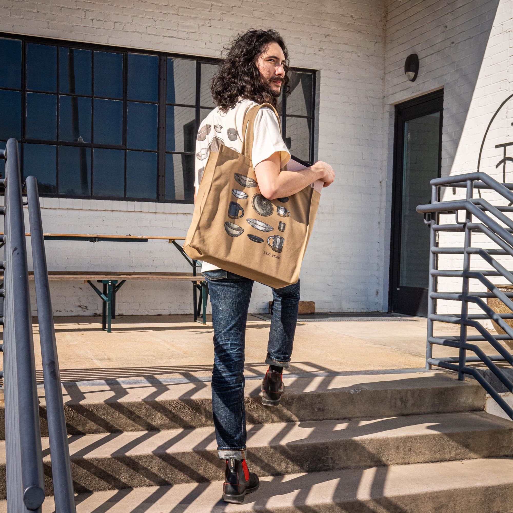 A person is standing on some steps about to enter a business wearing a heavyweight natural tote with an illustration of ceramic pottery on it